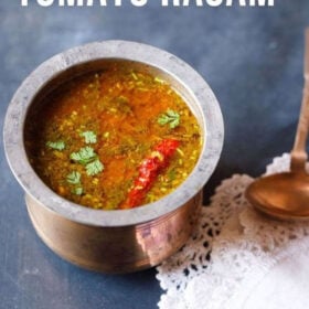 tomato rasam in a traditional South Indian container with a spoon placed on top of a white kitchen napkin on the left side on a blue board with text layovers.