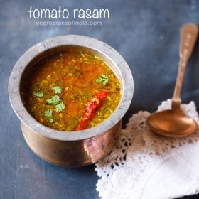 tomato rasam in a traditional South Indian container with a spoon placed on top of a white kitchen napkin on the left side on a blue board