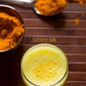 overhead shot of turmeric milk with ground turmeric kept in a bowl and steel spoon on a wooden table.