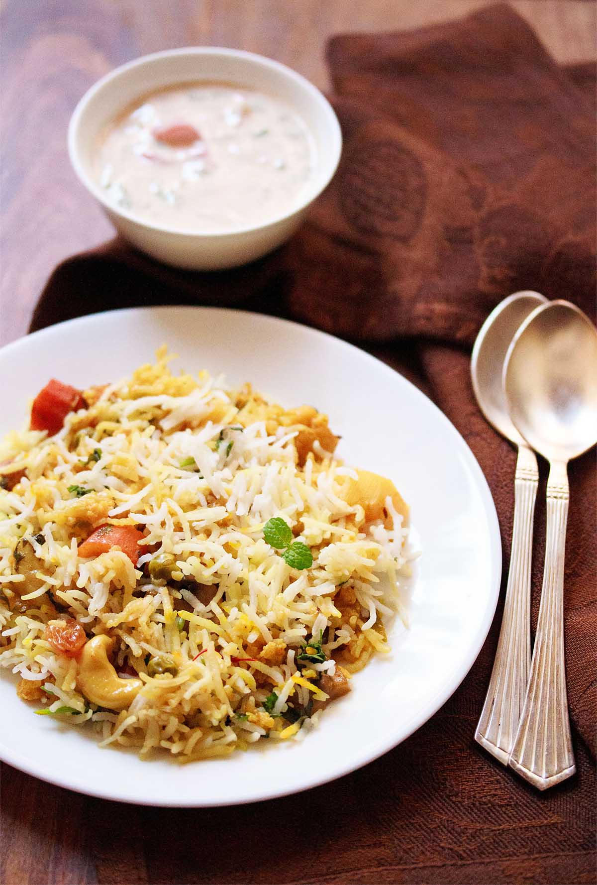 Top shot of veg biryani on white plate next to yogurt raita in small white bowl and two spoons on wooden table
