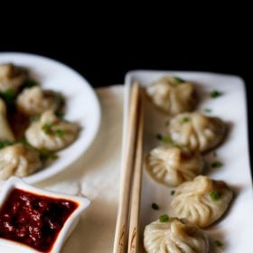 Veg momos arranged on a white rectangular long tray with a bamboo chopsticks by the side and a small square bowl of schezwan sauce at the side